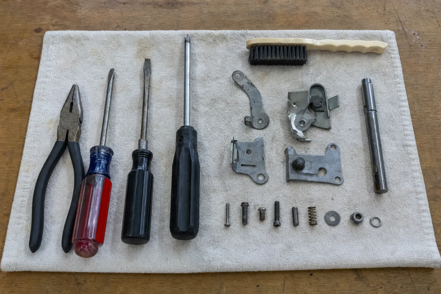 Hand tools and a dismantled appliance door latch laid out in order on a cloth over a workbench